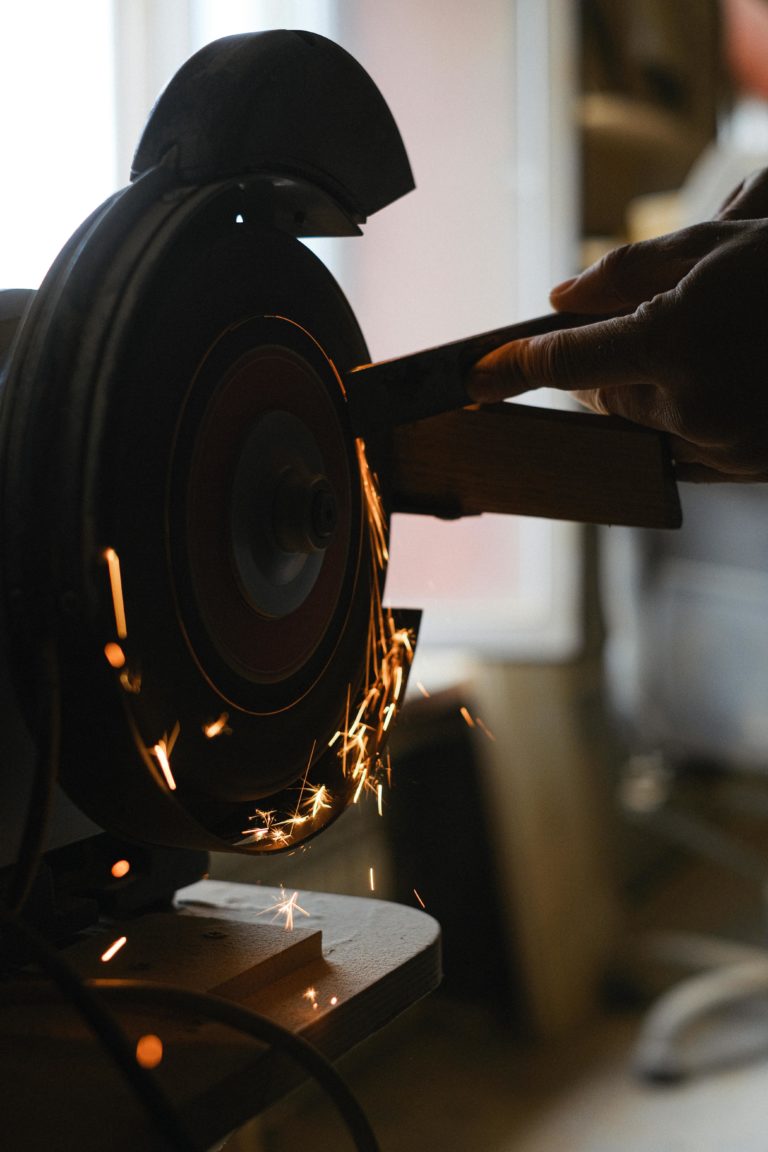 Unrecognizable craftsman using grinding machine to sharpen detail while standing in workshop with sparks on blurred background during work process