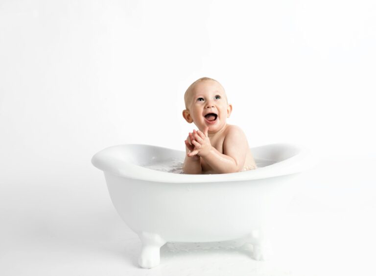 Cute baby laughing and clapping in a white bathtub against a white background.