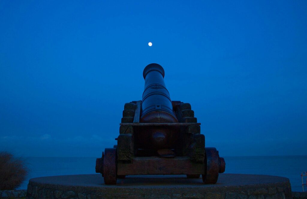 Silhouette of a historic cannon facing the moon in a serene midnight setting.