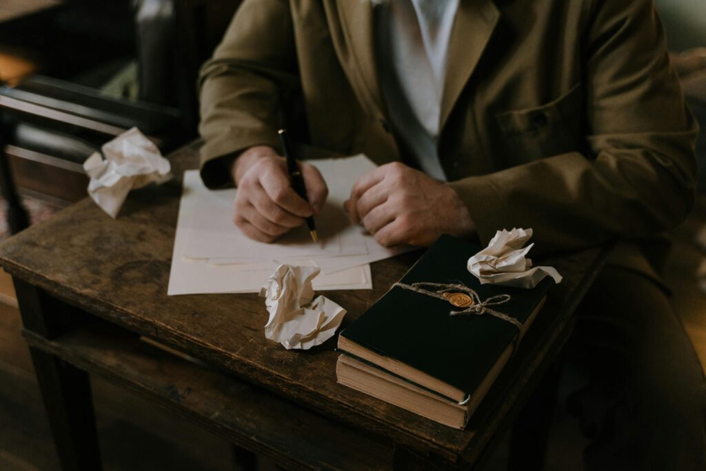 A person writing at a wooden desk with crumpled papers and a book.