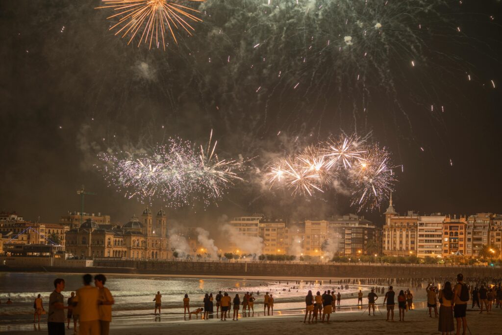 Vibrant fireworks illuminating the night sky over a coastal city with people watching from the beach.