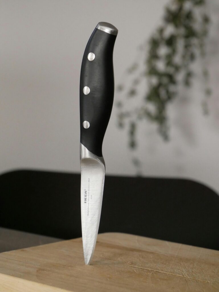 A stainless steel kitchen knife with a black handle standing on a wooden cutting board in a kitchen setting.